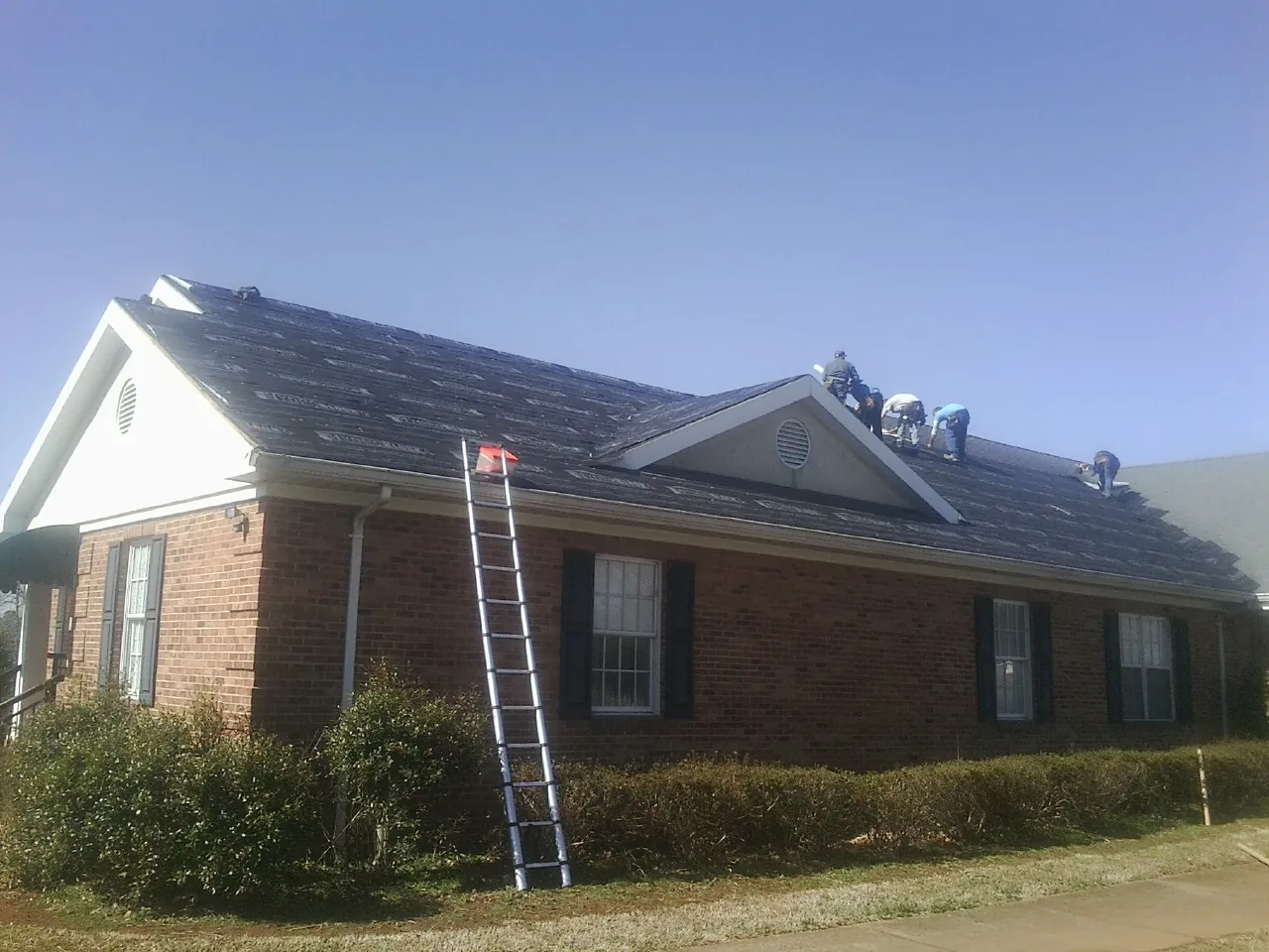 Crew tearing shingles off a brick ranch roof with a ladder set — Emergency Roof Repair work by our crew in Ossian