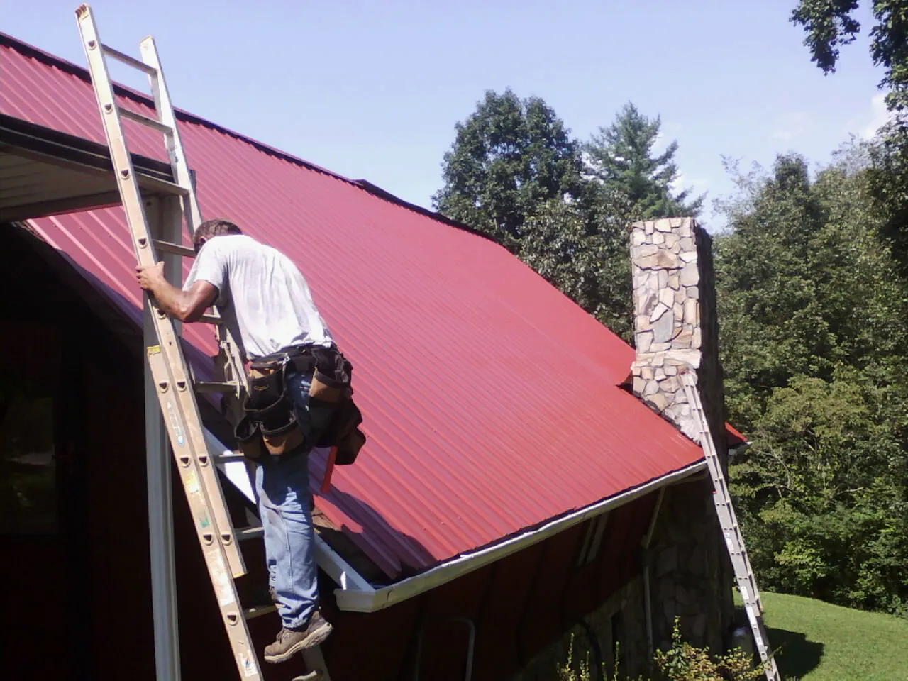 Roofer on a ladder beside a red metal roof and stone chimney — Soffit & Fascia Repair by Roofing Brothers in Ossian